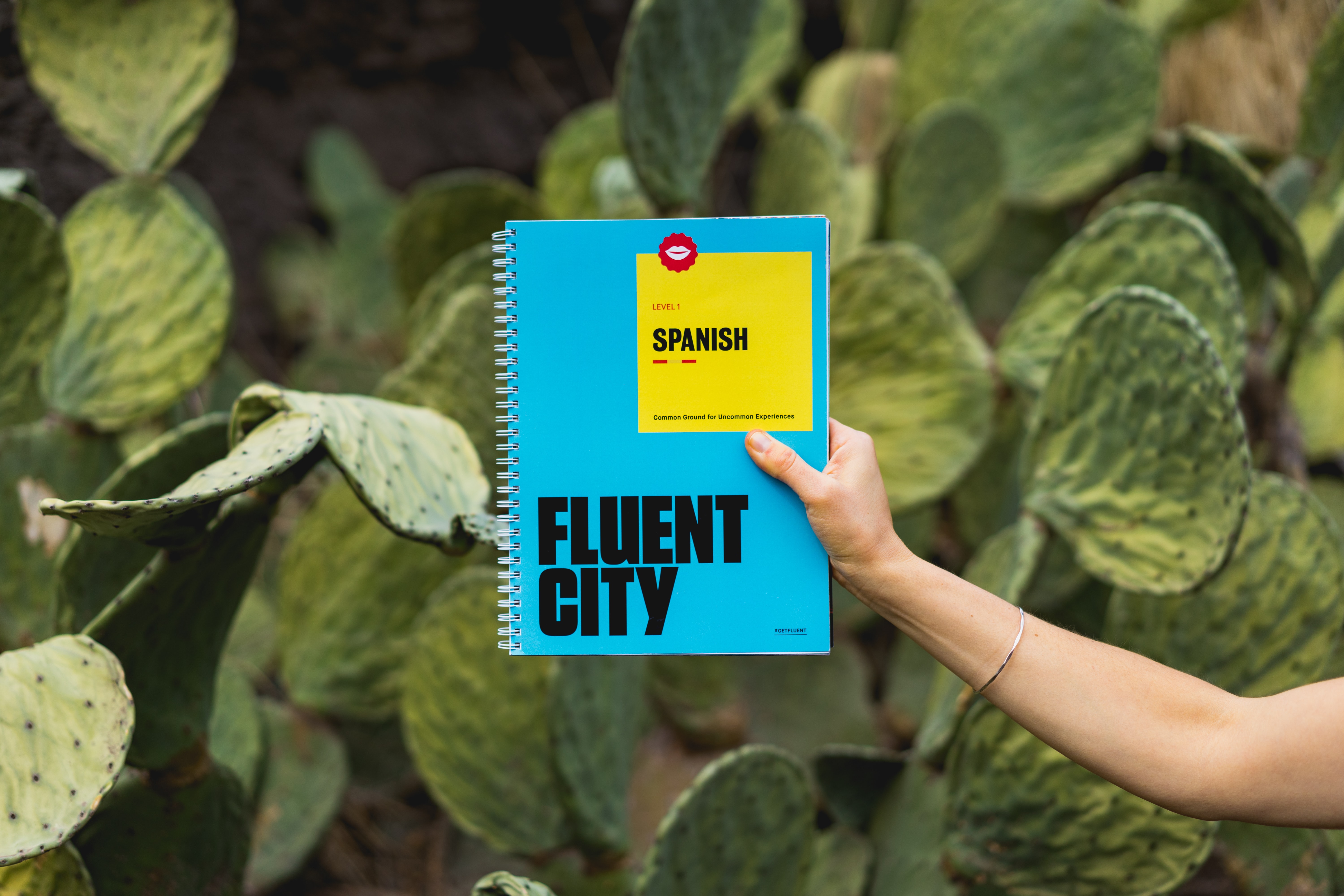 Hand holding notebook reading "Fluent City: Spanish". Cactus background.